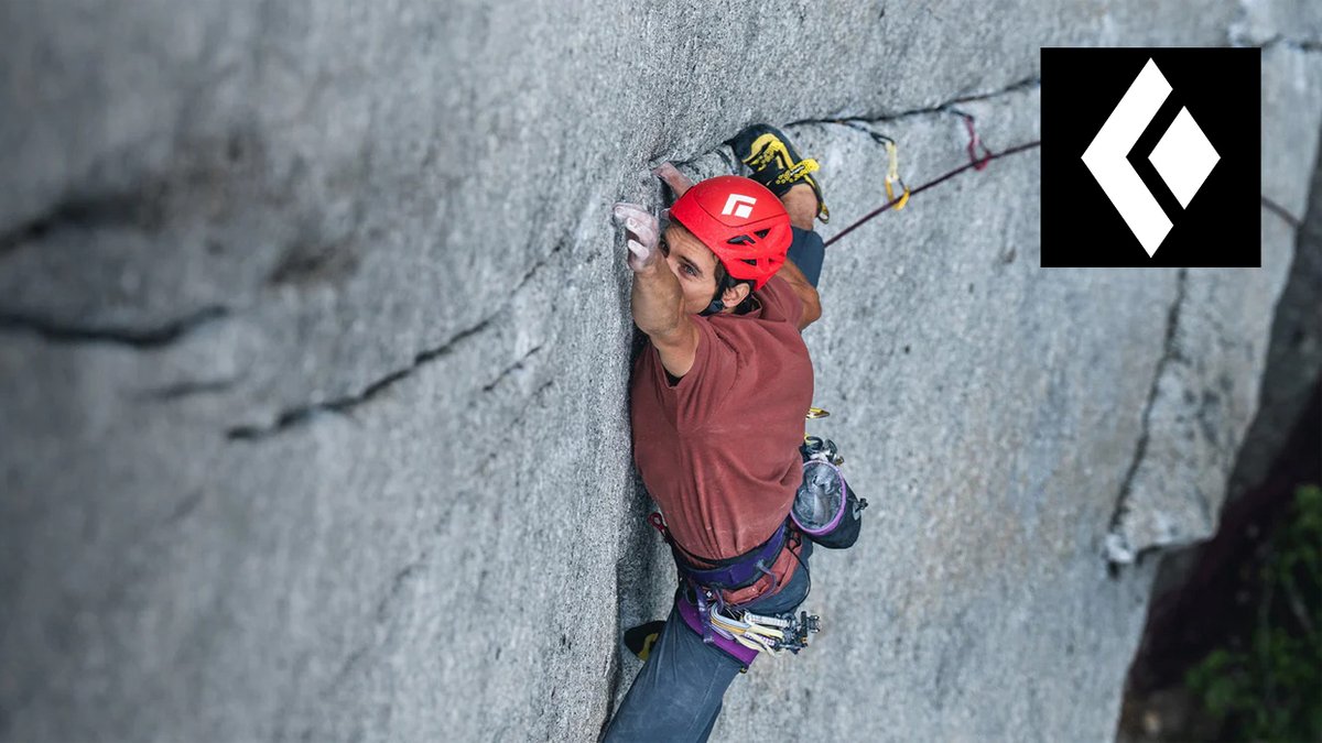 A rock climber wearing a red Black Diamond helmet and climbing harness scales a vertical grey cliff face.