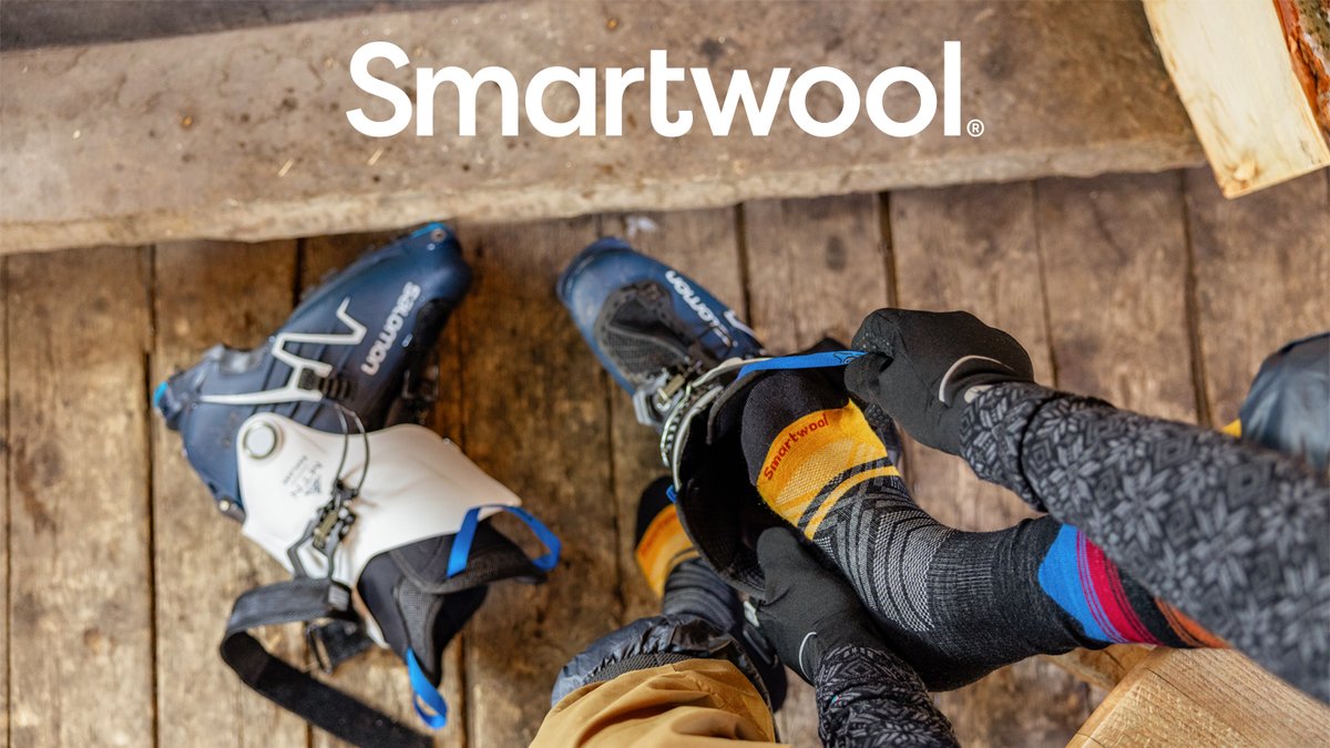 A first-person view of a person putting on colorful Smartwool socks and Salomon ski boots on a wooden floor.
