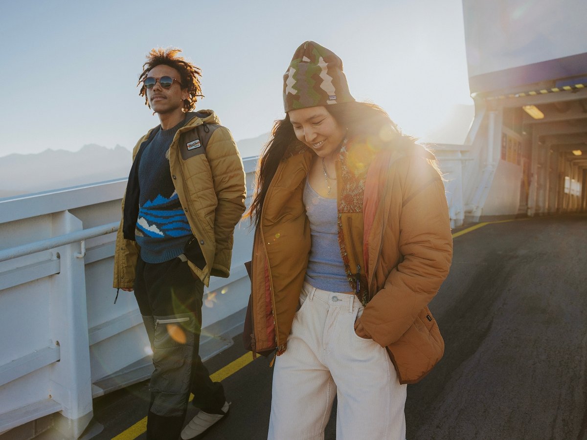 A man and a woman wearing big fall jackets stand on the sunlit deck of a ferry.