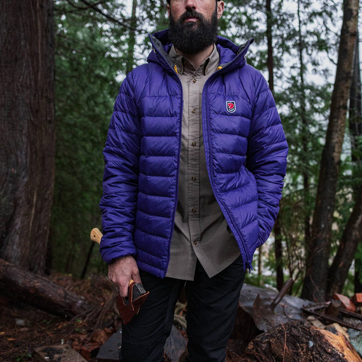 A man wears a Fjallraven Expedition jacket, standing in the woods while holding a hatchet.