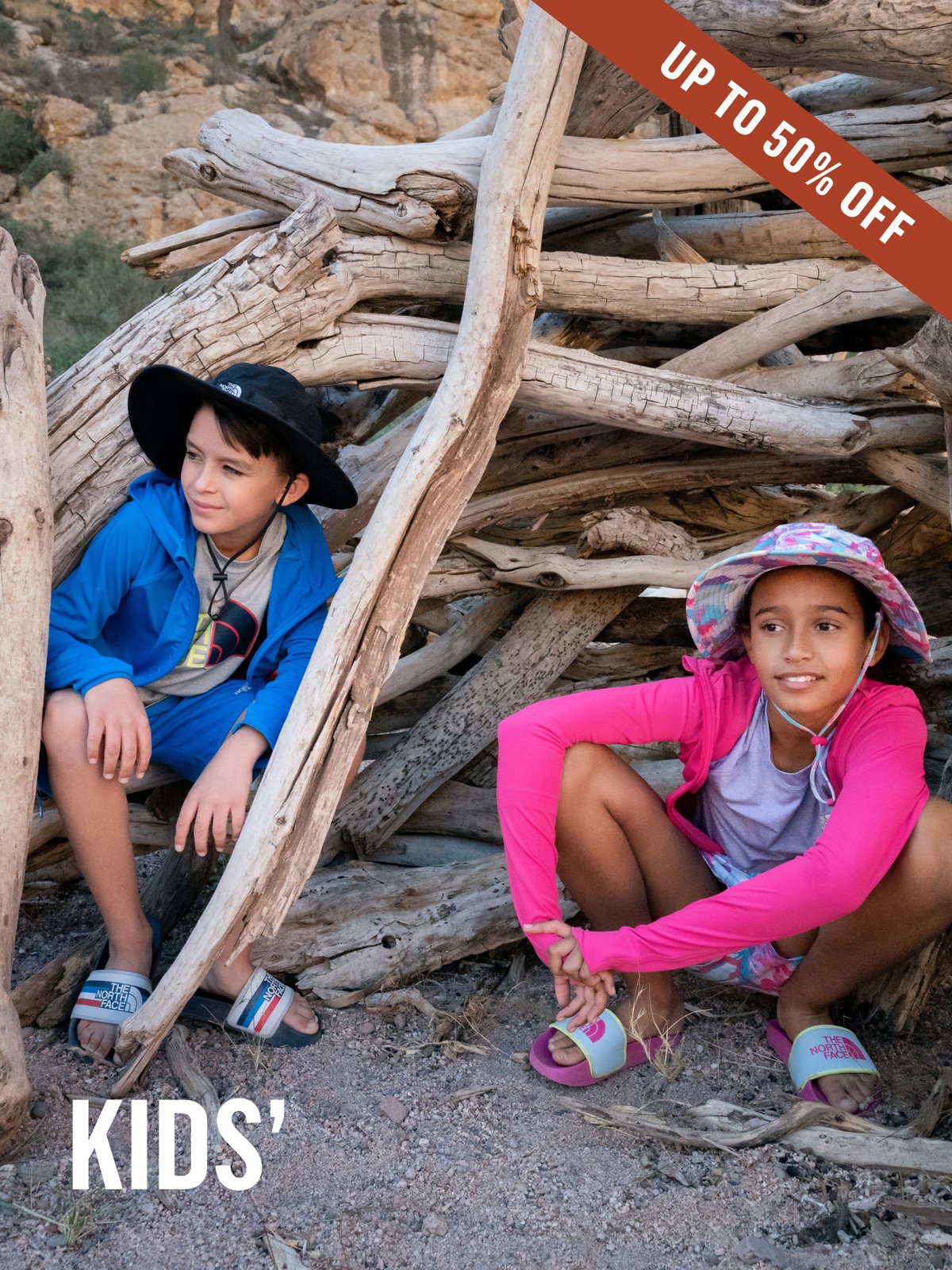 Two kids crouch inside a driftwood fort in a desert canyon, wearing The North Face sun shirts, bucket hats, and slide sandals. Kids' Up To 50% off.