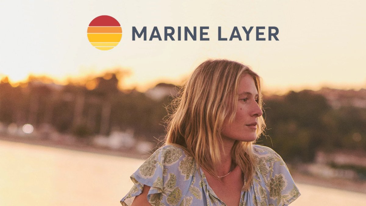 A woman with reddish-blonde hair gazes toward the horizon during golden hour, wearing a patterned blue shirt with the Marine Layer logo and striped sunset graphic visible above her.