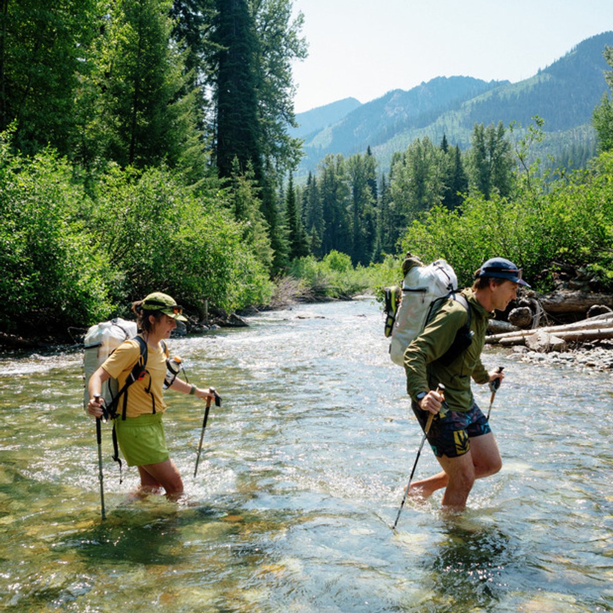 Two hikers wade through a river, wearing packs on their back and Sunblocker tops.