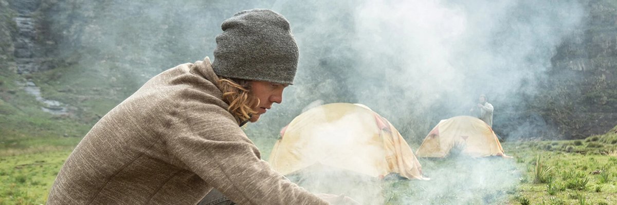 Woman cooking over a campfire in a mountain valley while camping with tents in the background.