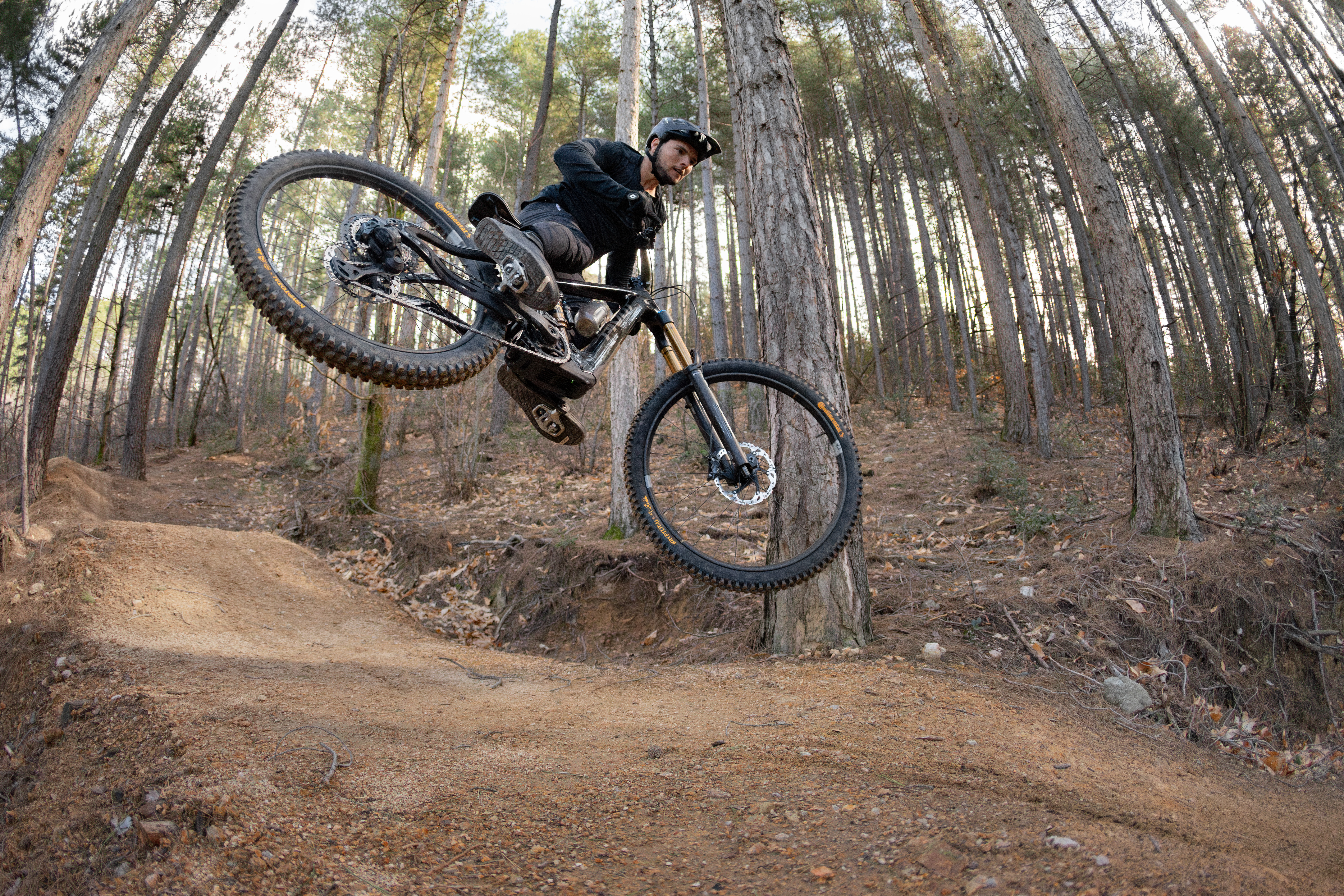 Rider whipping full-suspension eMTB off dirt jump wearing all-black MTB kit and helmet on dry forest trail in autumn conditions