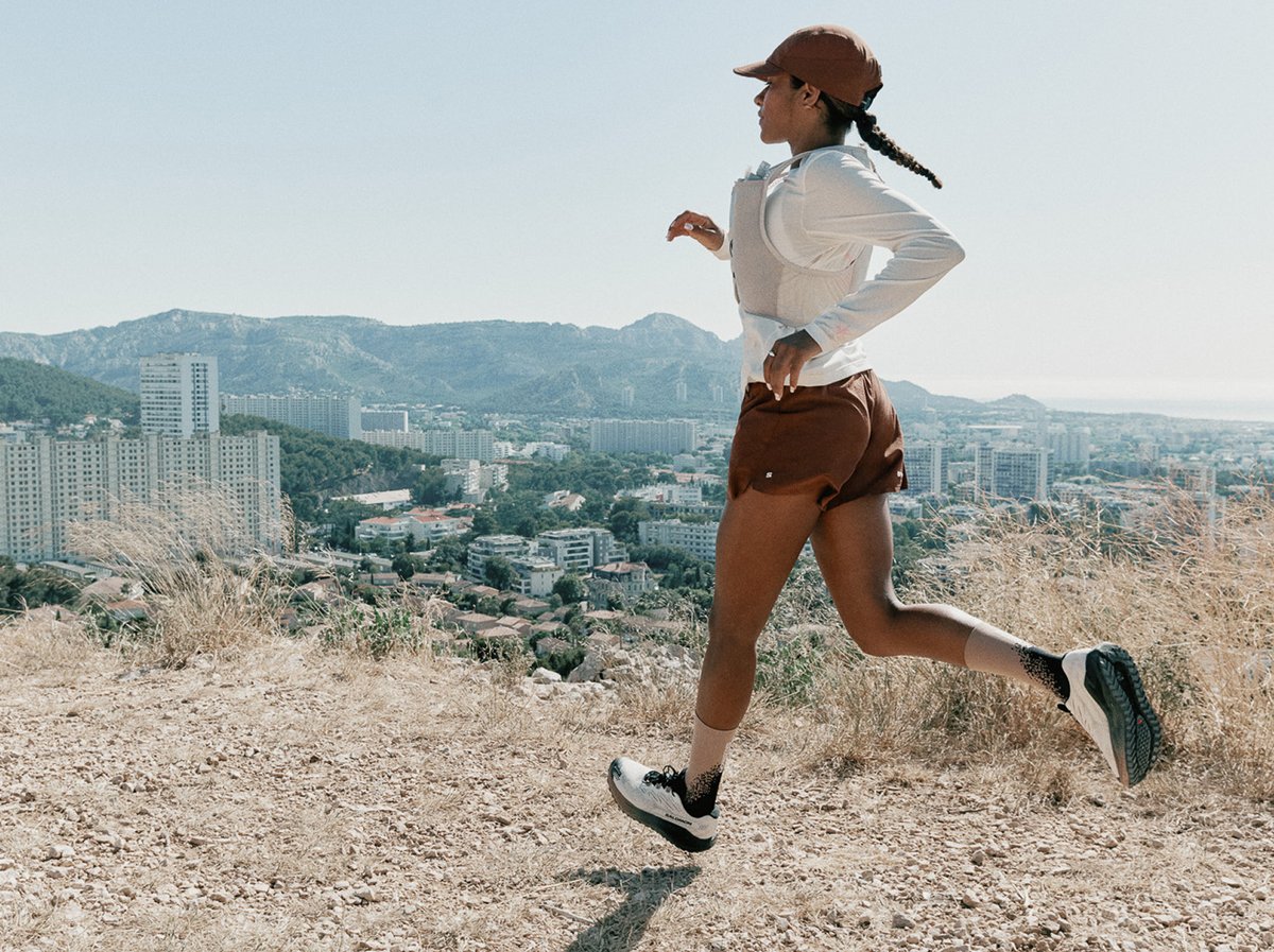A runner in a white long-sleeve and rust shorts sprints a dry hillside trail above a sun-baked city skyline, mountains behind.