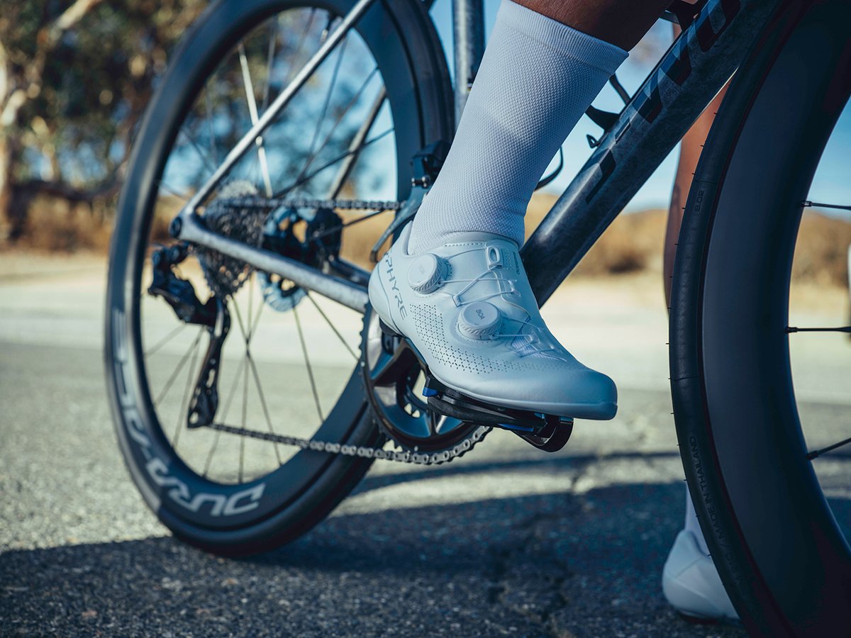 Side profile of a white S-PHYRE shoe with dual BOA dials, standing next to a Dura-Ace-branded wheel, desert road backdrop