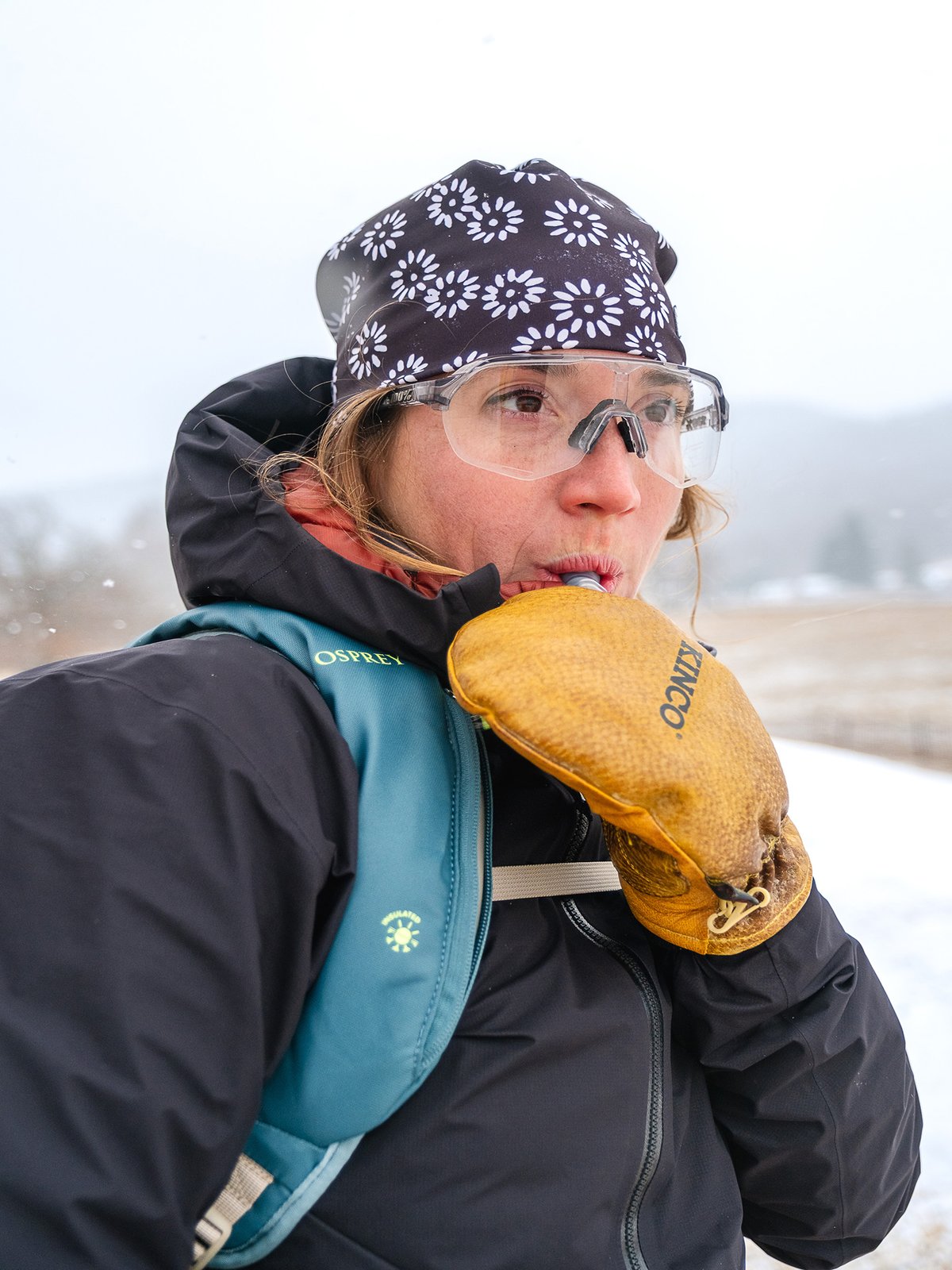 A woman wearing goggles drinks from a hydration system.