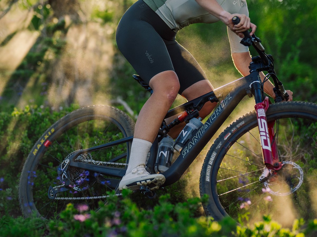 Rider on a Santa Cruz mountain bike with RockShox fork and Maxxis tires rolls through a sun-dappled trail lined with wildflowers.