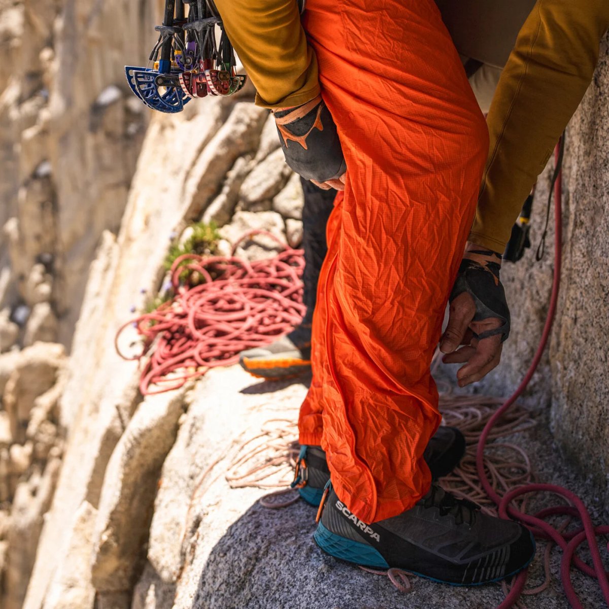 Climber wearing technical climbing pants with spiral zip cuffs over approach shoes standing on granite ledge during trad climb