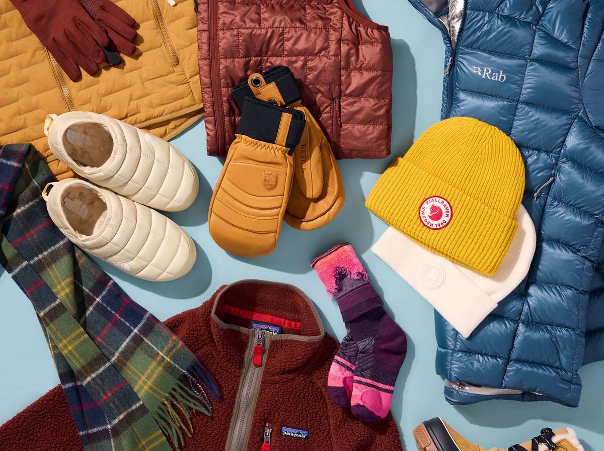 A flat lay of winter accessories and clothing on a light blue background includes beanies, mittens, ski socks, insulated slippers, leather glvoes, a plaid scarf, a fleece quarter-zip, and puffy jackets.