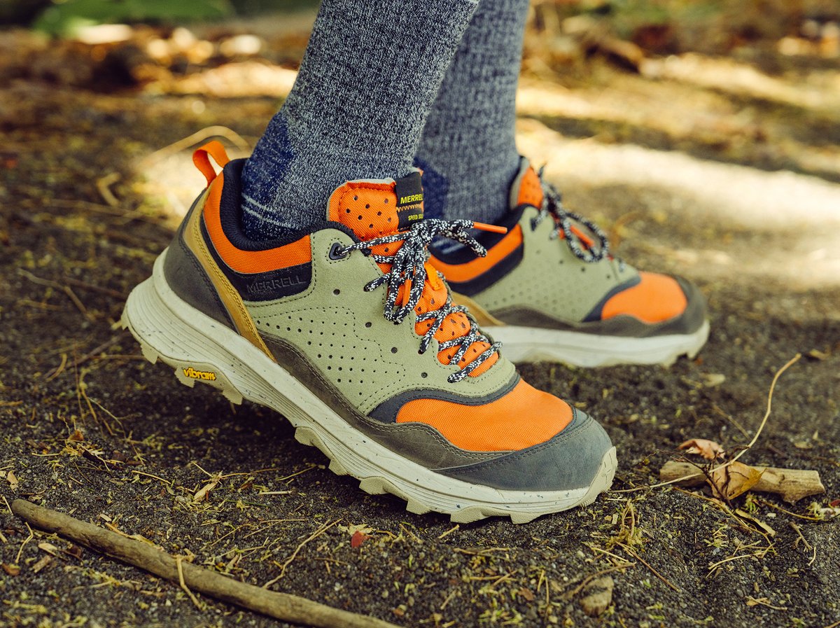 A close-up view of Merrell hiking shoes, which show the yellow Vibram logo on the side of the outsole, worn by a person who stands on a dirt trail surrounded by fallen leaves and sticks.