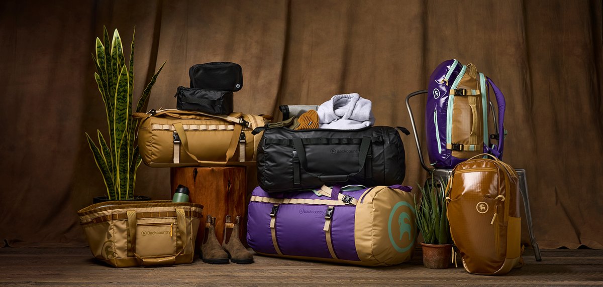 Backcountry duffel bags in tan, black, and purple arranged in a studio setting with plants and gear.
