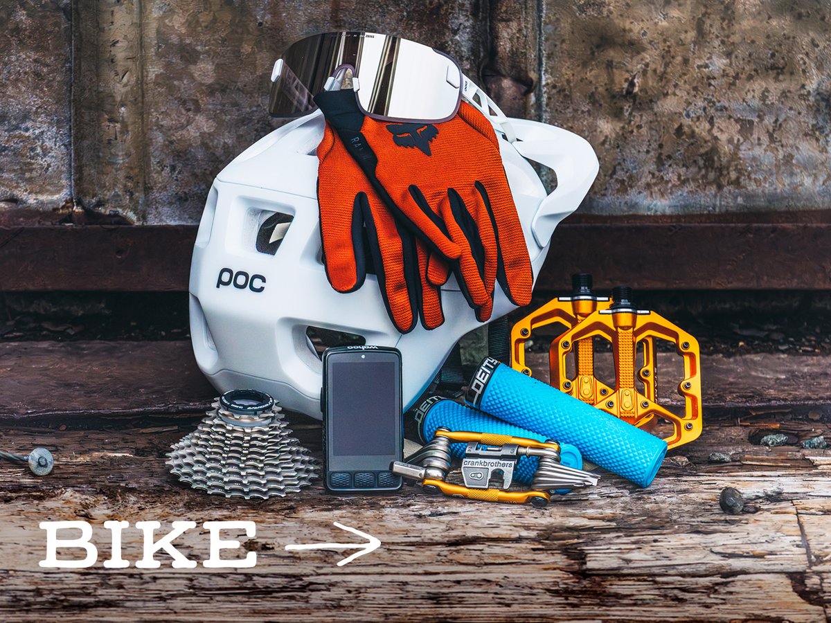 Mountain bike gear, including a helmet, gloves, glasses, flat pedals, grips, a cassette, a multitool, and a bike computer, on weathered wood: “Bike.”