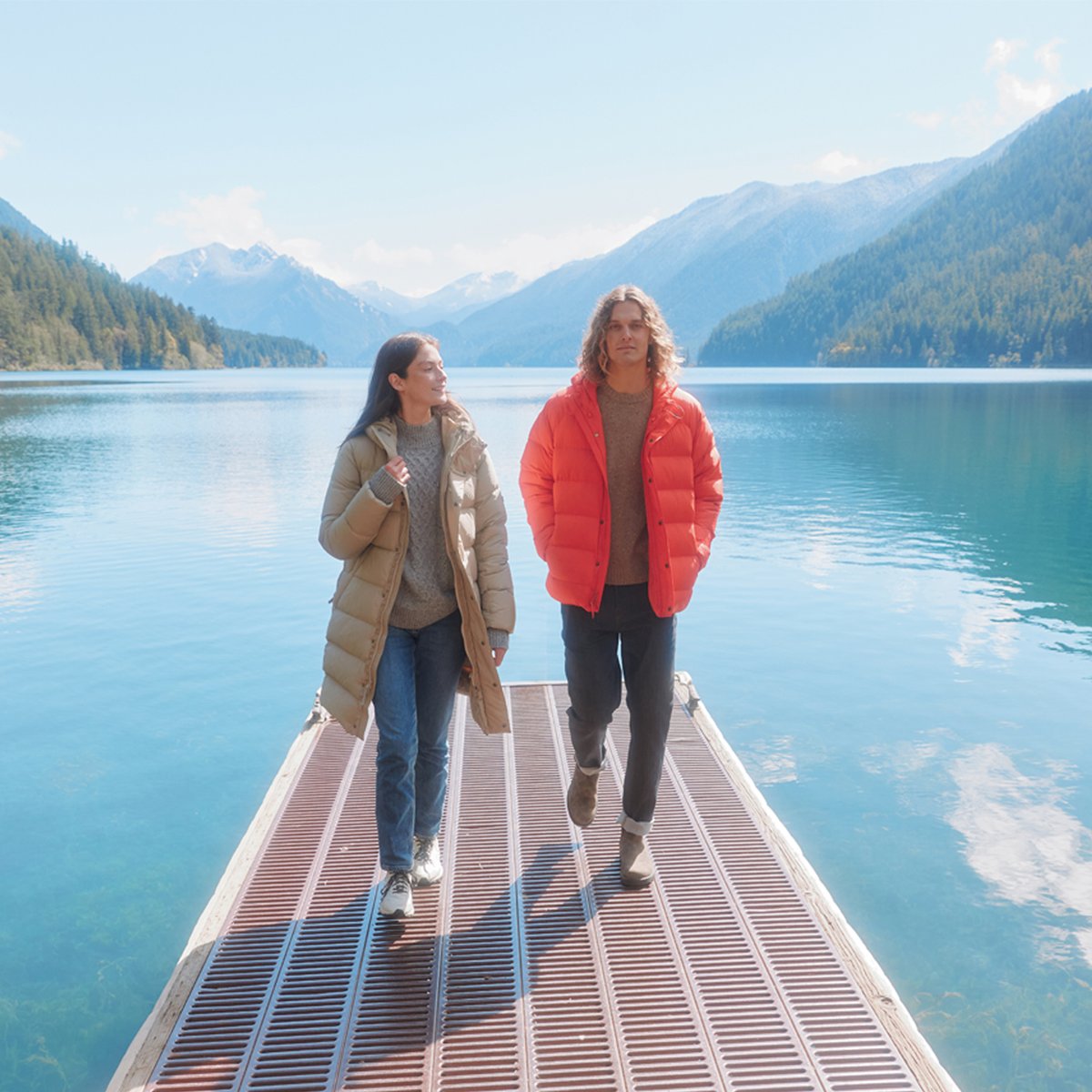 A man and a woman wearing puffers and Stretch Zion pants walk along a pier, the sky reflected in the clear lake near them.