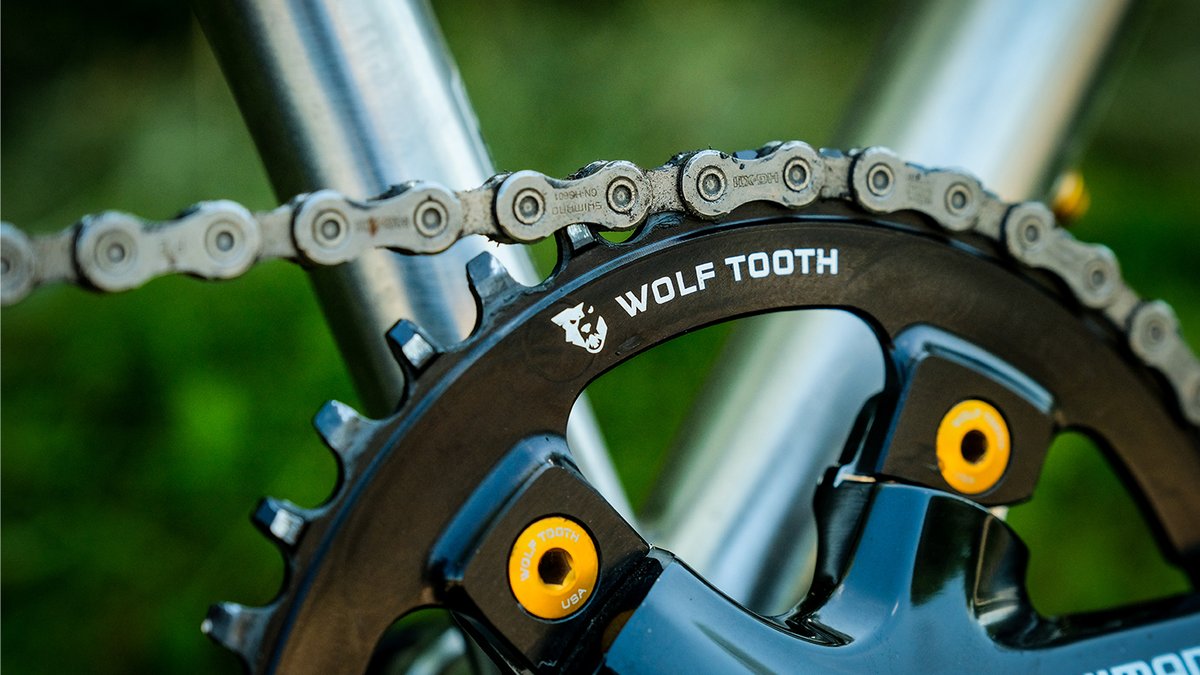 Close-up of a Wolf Tooth crankset with silver chain and yellow brake calipers mounted on a blue bicycle frame against a green blurred background.