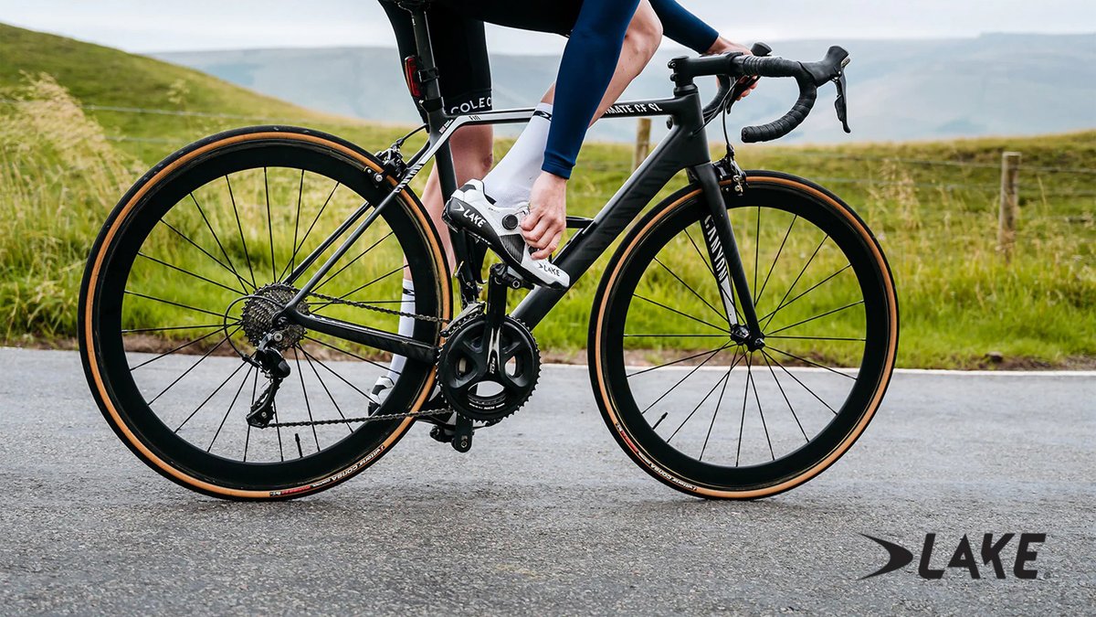 A cyclist in blue leg wear and white shoes rides a black road bike with distinctive orange-rimmed wheels on a scenic rural road with green hills in the background.