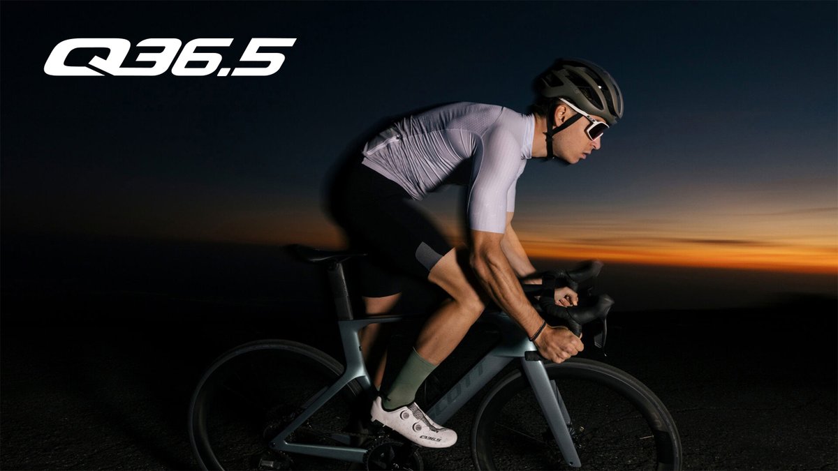 Professional cyclist riding a Q35.5 road bike at dusk wearing aerodynamic cycling gear, helmet, and sunglasses on a dark road with sunset sky backdrop.
