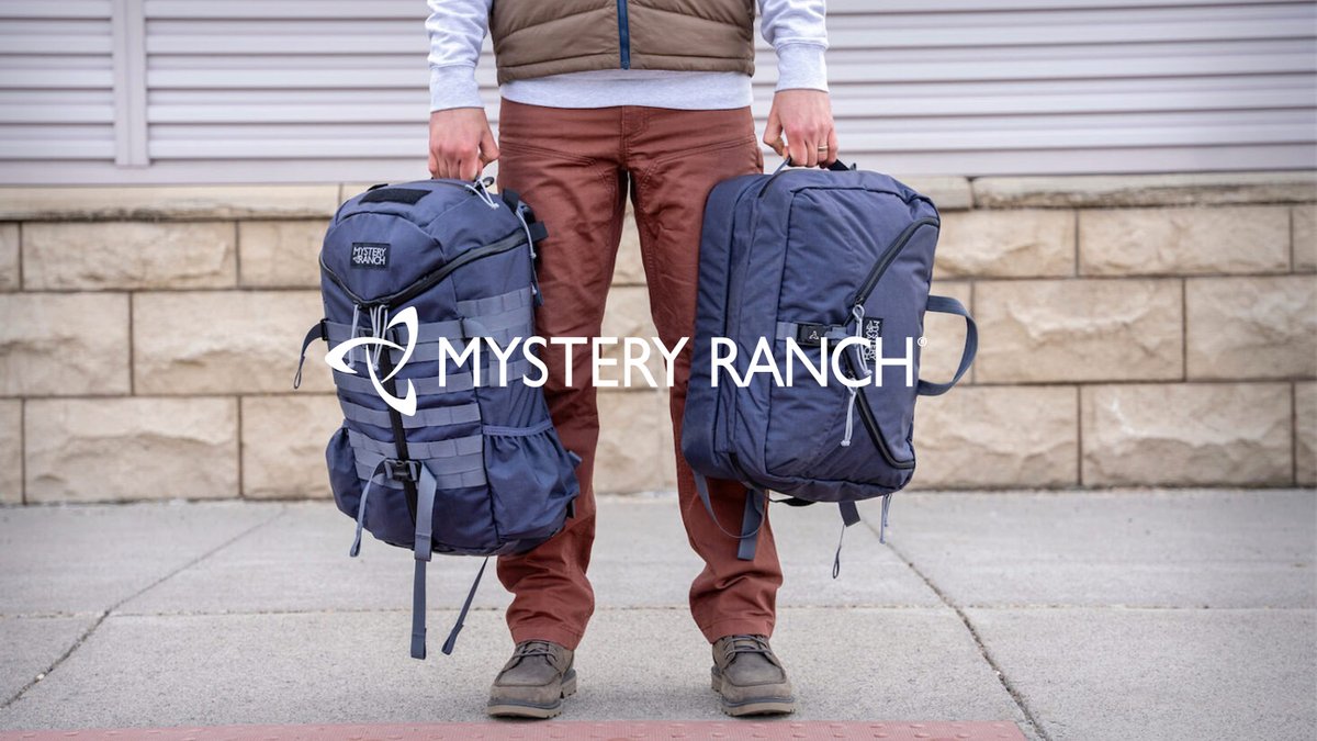 Person holding two navy blue Mystery Ranch backpacks while standing outdoors wearing casual hiking attire.