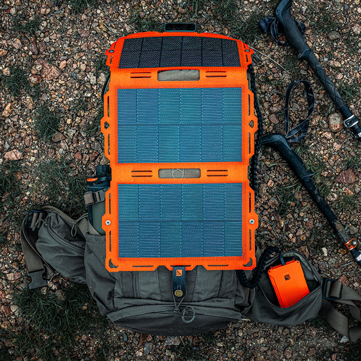 An orange Dark Energy Spectre folding solar panel unfolded across the top of a backpack on rocky ground, with trekking poles and an orange Poseidon power bank beside it.