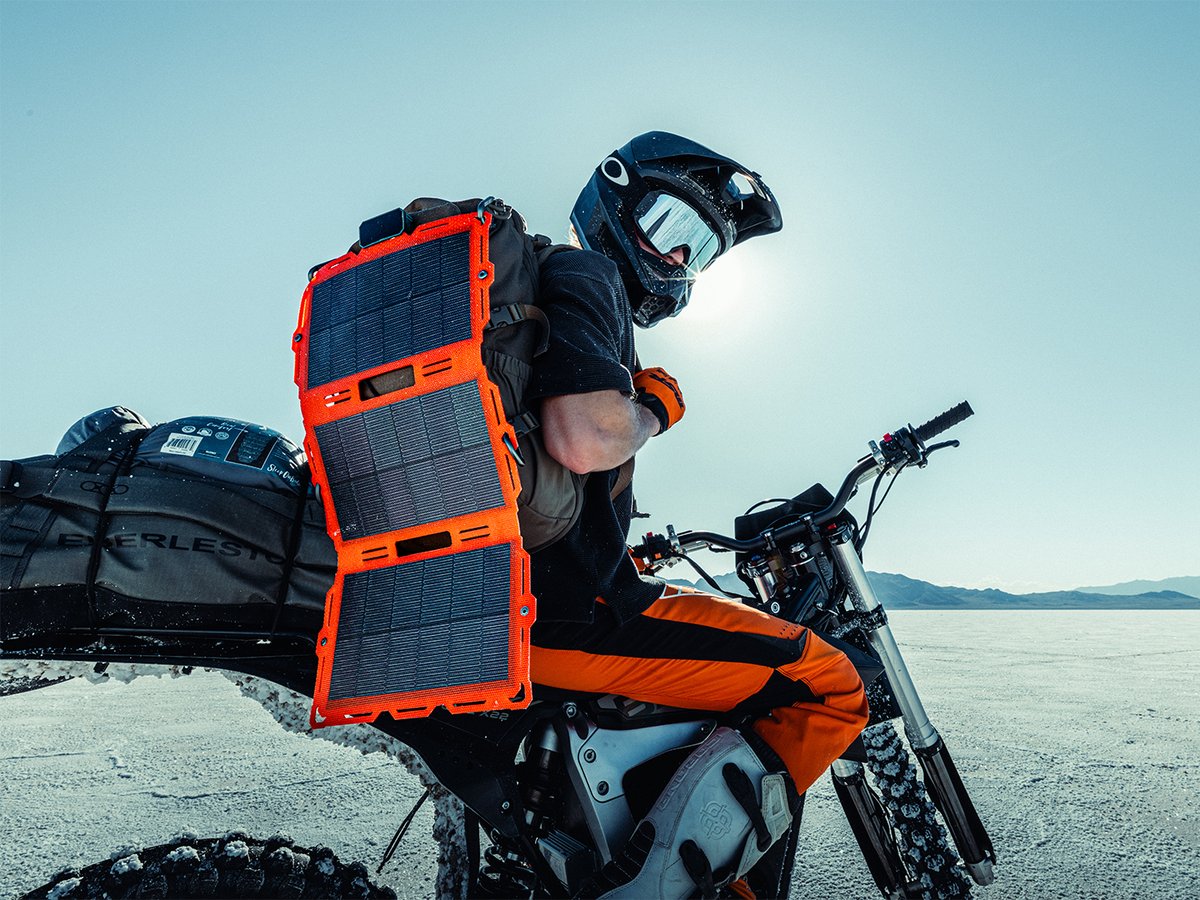 A dirt bike rider stops on the salt flats with an orange Dark Energy Spectre folding solar panel strapped to a backpack.