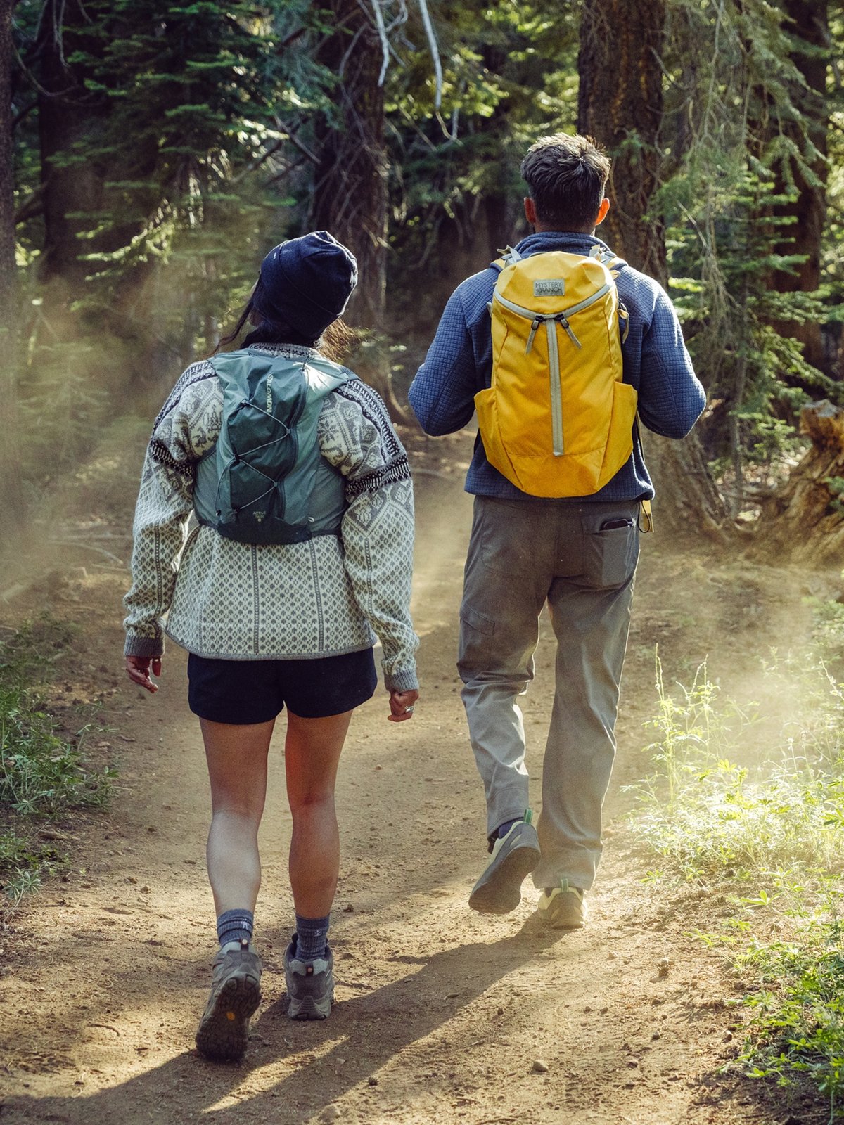 Two hikers on a dirt trail in sunlit forest, both carrying backpacks.
