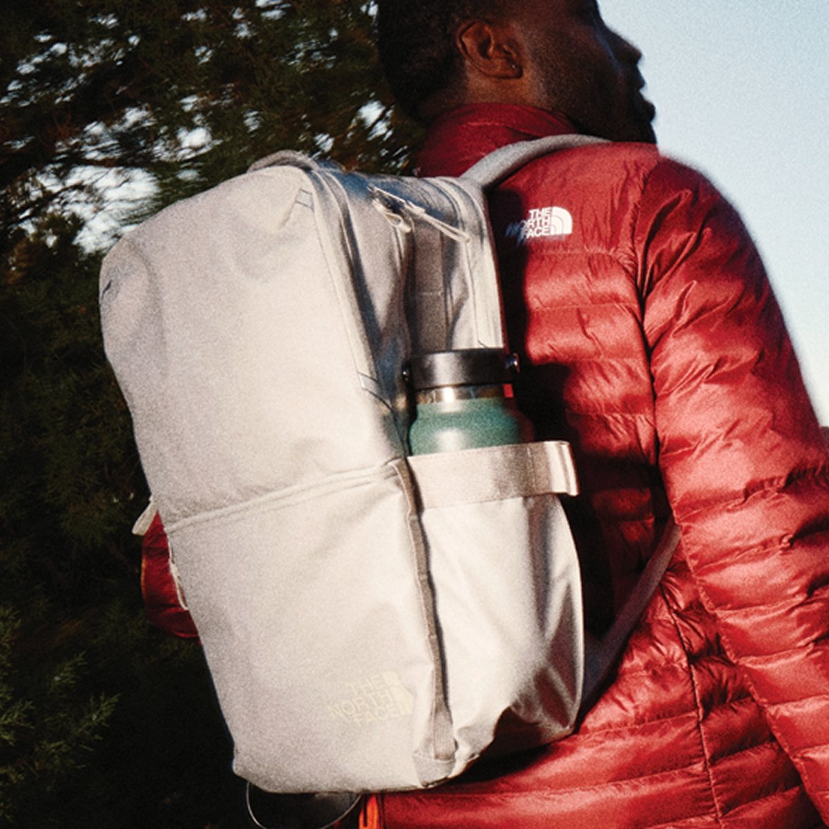 A person in a red puffer jacket carries a light gray backpack with a water bottle.