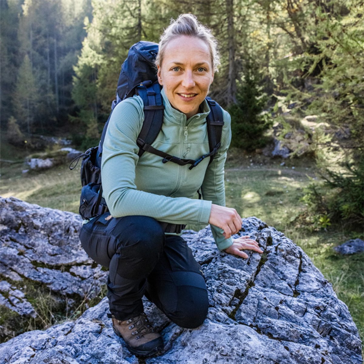 Woman wearing a blue hiking backpack and green jacket crouching on a rock in a forest with a stream in the background.