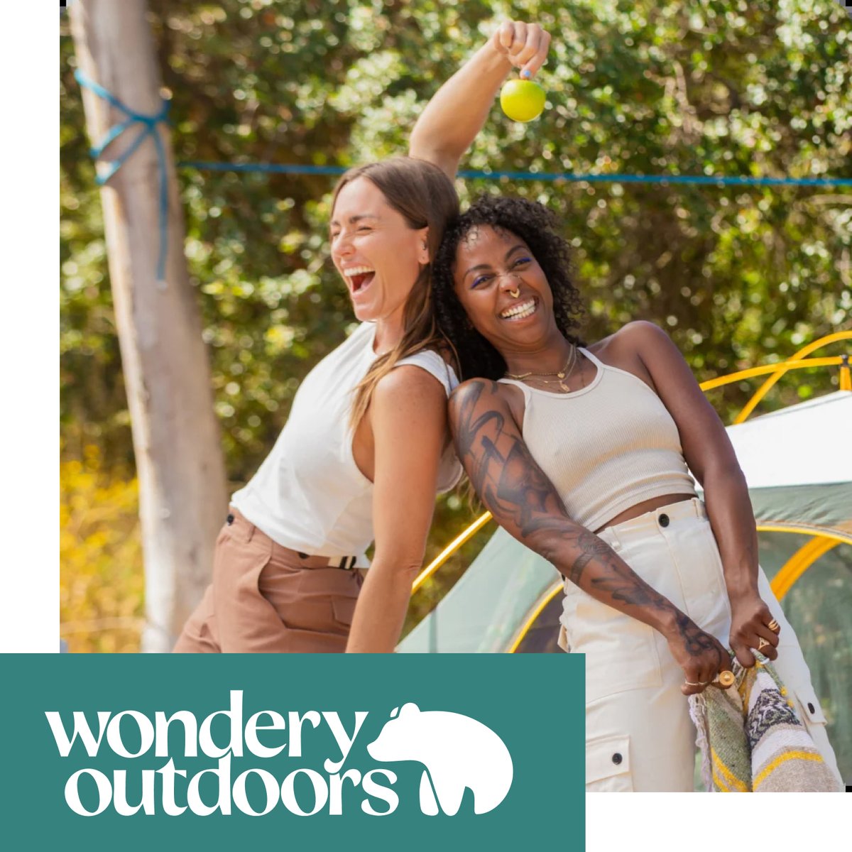 Two women celebrating outdoors, tossing a ball in a festive camp scene. Shop Wondery outdoors.