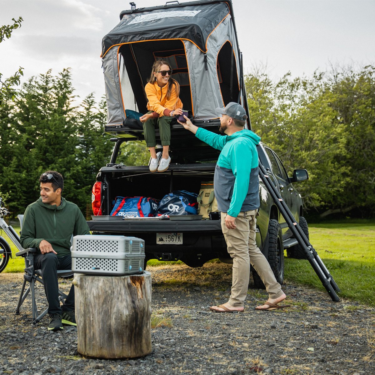 A group of friends sits at a campsite, wearing Ferrosi gear. A woman in a rooftop tent waves down to her friend on the ground.