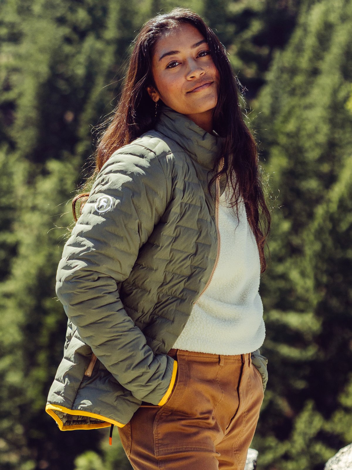 A woman with long hair wearing a green quilted jacket, white sweater, and brown pants stands in a pine forest.