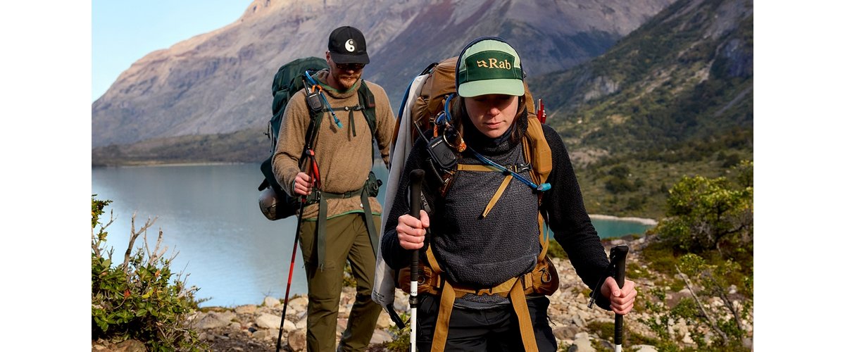 Two people hike on a rocky mountain trail, each wearing outdoor gear and carrying large backpacks and trekking poles. The landscape features a lake, green slopes, and rugged mountain peaks in the background beneath a clear sky. The person in front wears a green "Rab" cap.
