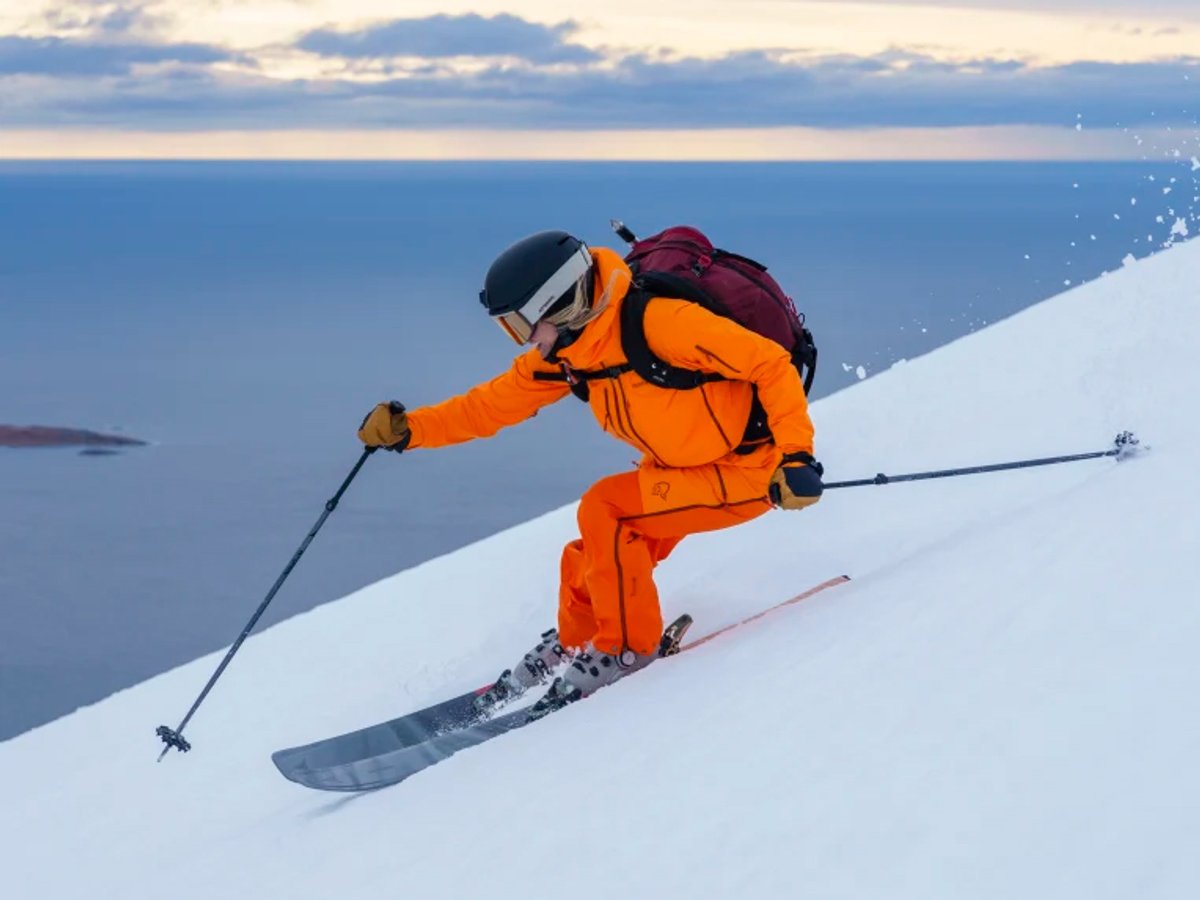 Professional skier in bright orange jacket and helmet descending snowy mountain slope with ocean and blue sky in background.
