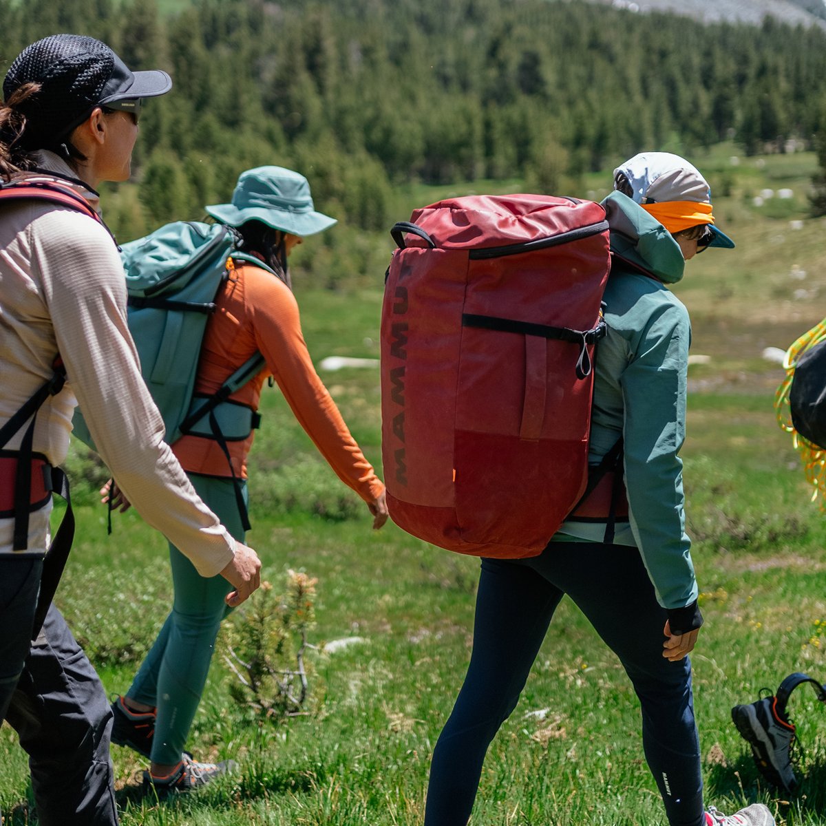 Three people with backpacks hike through a grassy field.