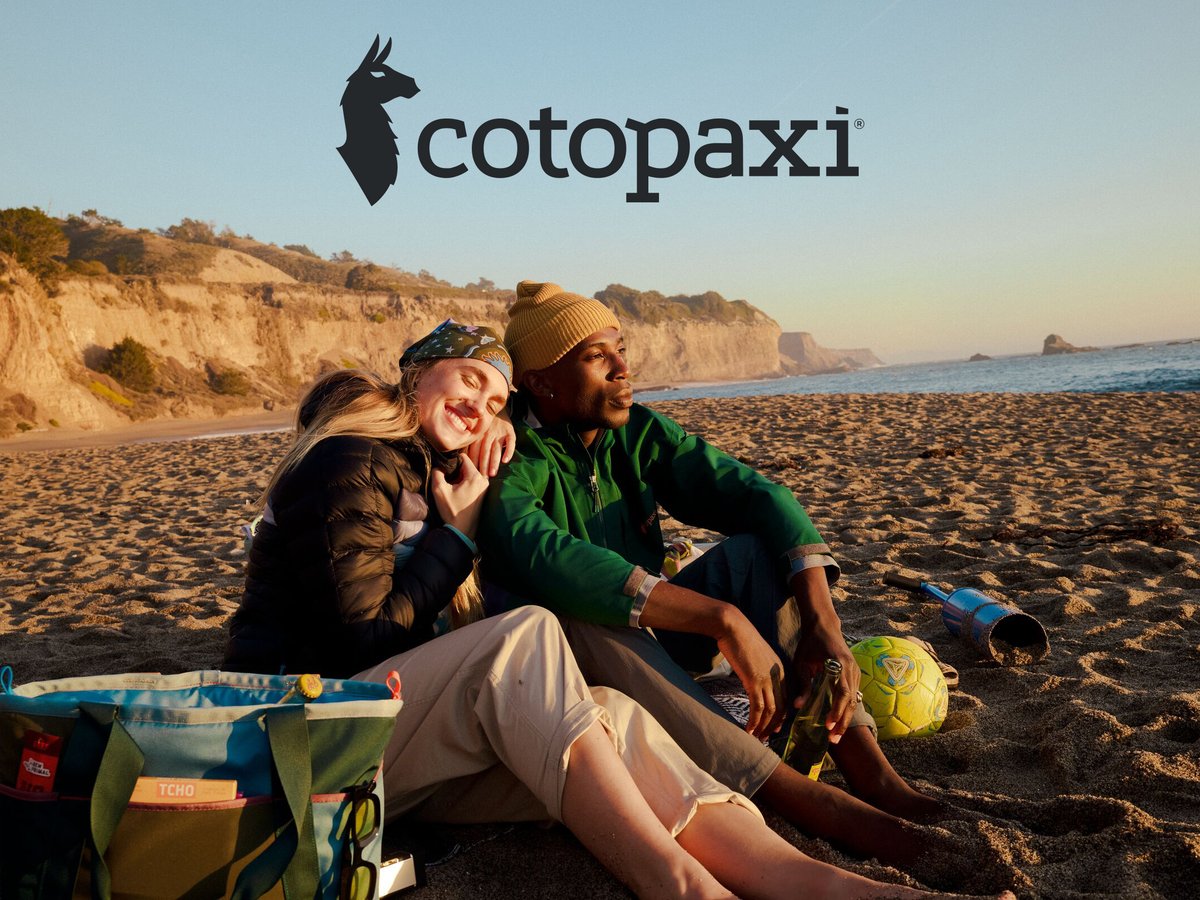 Two friends sitting on a beach at sunset next to a Cotopaxi cooler bag; the Cotopaxi logo is centered at the top.