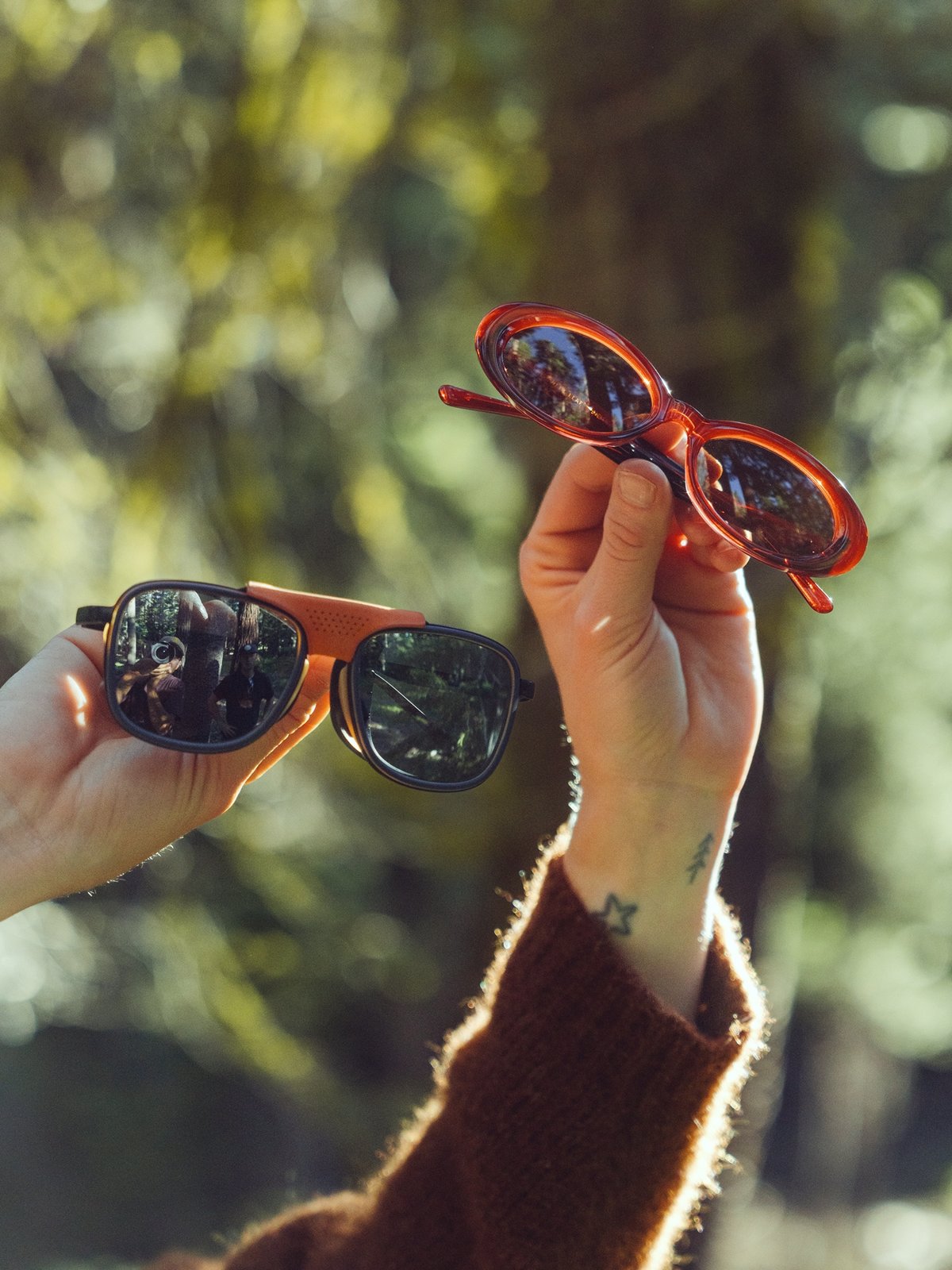 A hand with a small star tattoo on the wrist holds up a pair of round orange sunglasses outdoors, with a background of trees and sunlight.