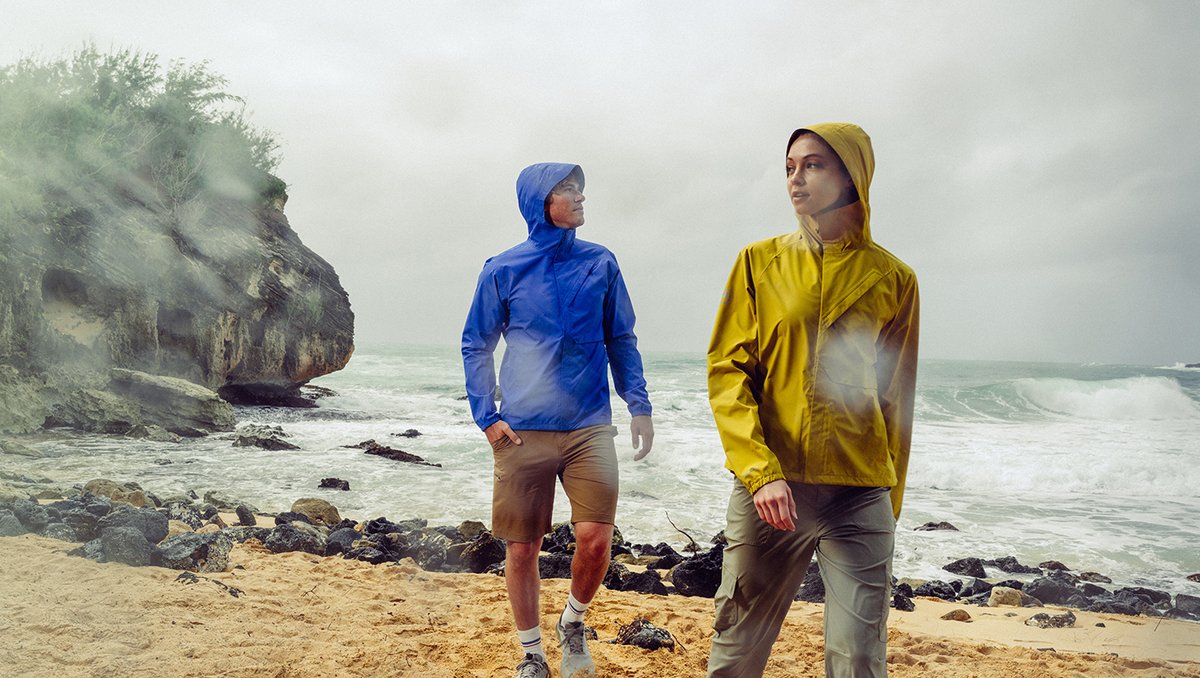 Two hikers walking windswept rocky shoreline in hooded waterproof jackets with crashing ocean waves and sea cliffs behind them