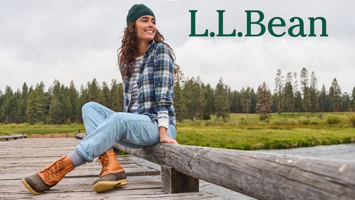 A woman wearing a plaid shirt, beanie, and Bean Boots sits on a wooden bridge overlooking a river and pine forest.