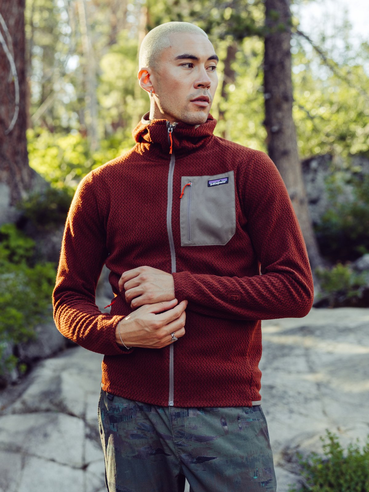 A man in a forested area wearing a textured red zip-up jacket with a gray chest pocket and gray camouflage pants.