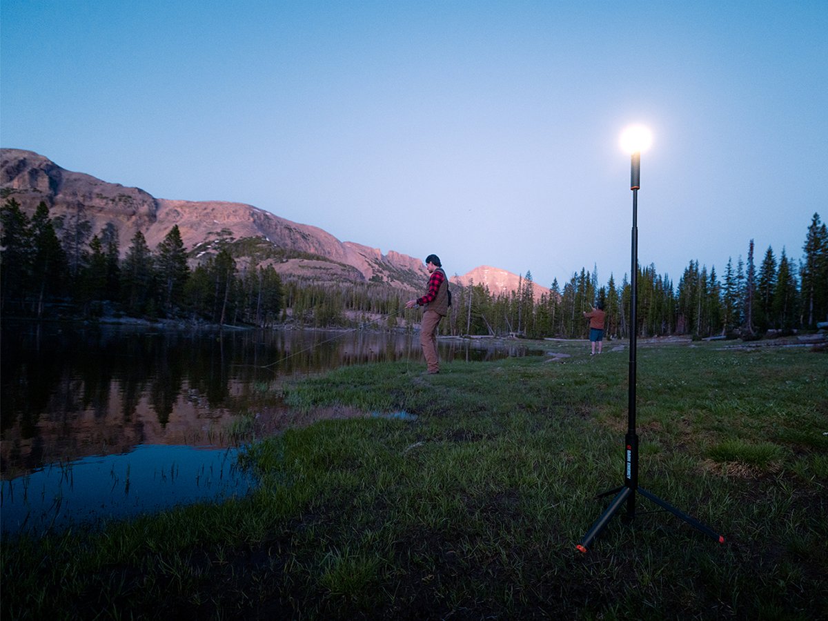 A Dark Energy Sentinel telescoping light stands on its tripod base at dusk, lighting a grassy lakeshore while two anglers fly fish, with alpenglow on the peaks behind.