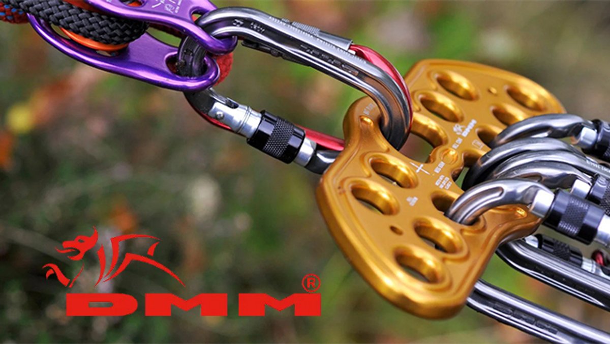 A close-up of climbing carabiners and belay devices in purple and gold with the DMM brand logo visible against a blurred outdoor background.