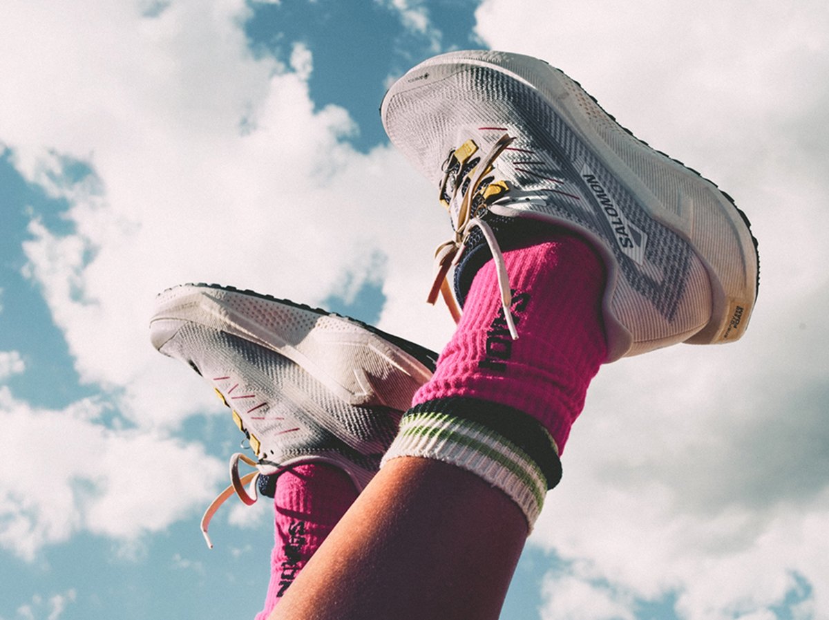 A pair of legs wearing white Salomon running shoes and bright pink socks with green and white stripes held up in the air, seen before the scattered clouds and blue sky as if their person is laying on their back. 