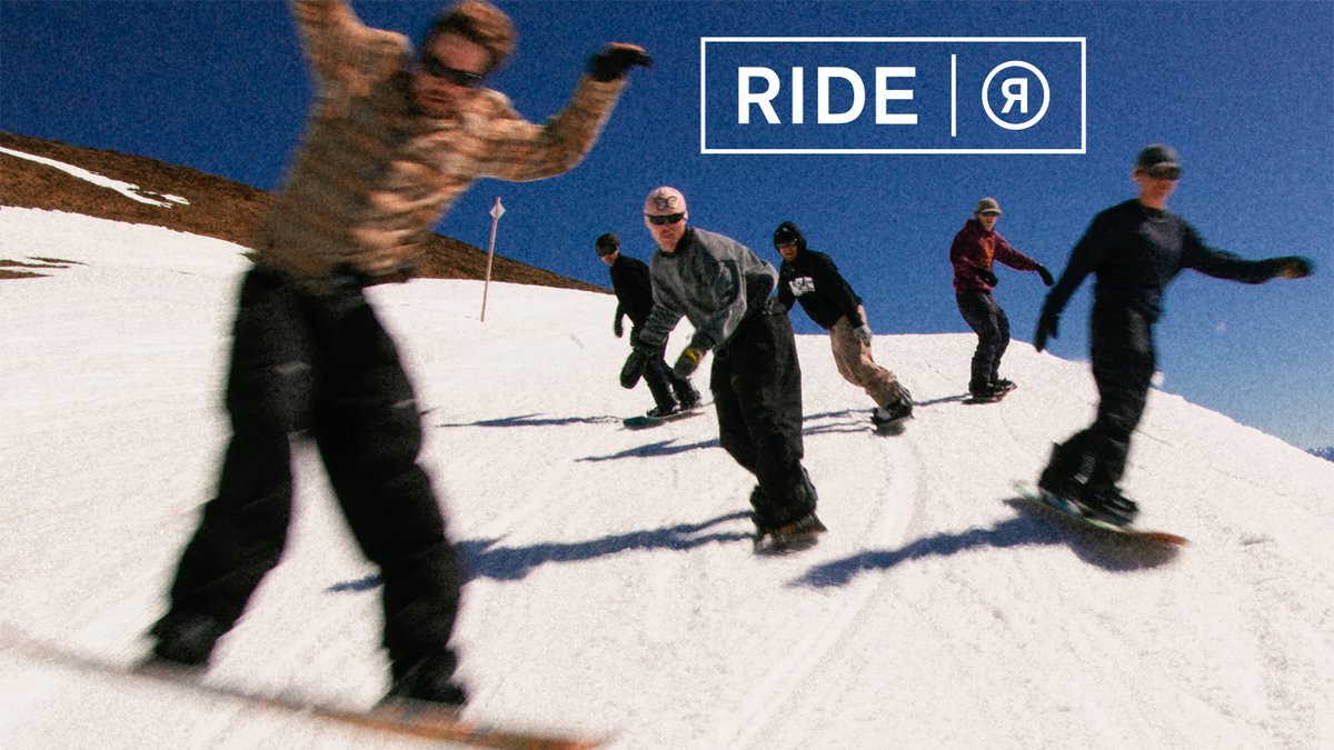 Group of snowboarders riding down a snowy mountain slope on a clear blue sky day with the RIDE brand logo displayed.