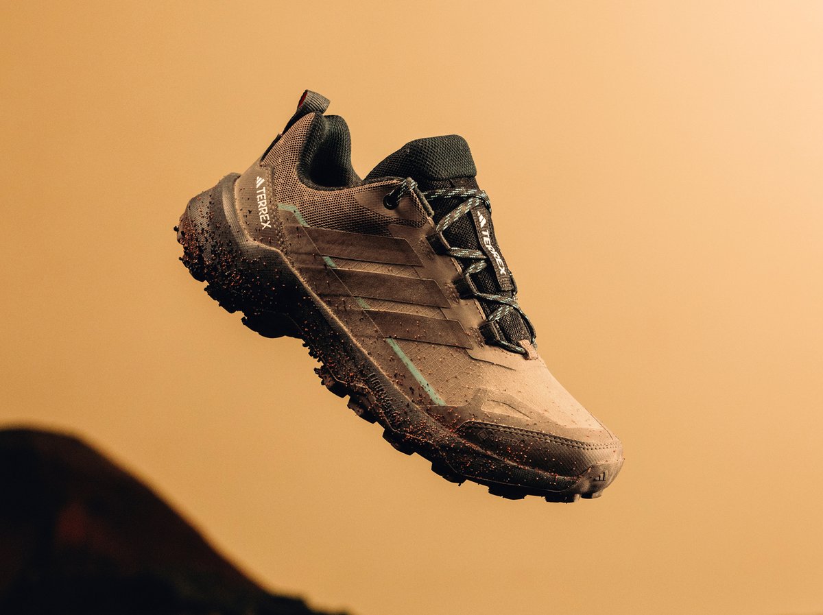 A mud-splattered brown and black trail running shoe with the word "TERREX" on the side is suspended in mid-air against a plain tan background.