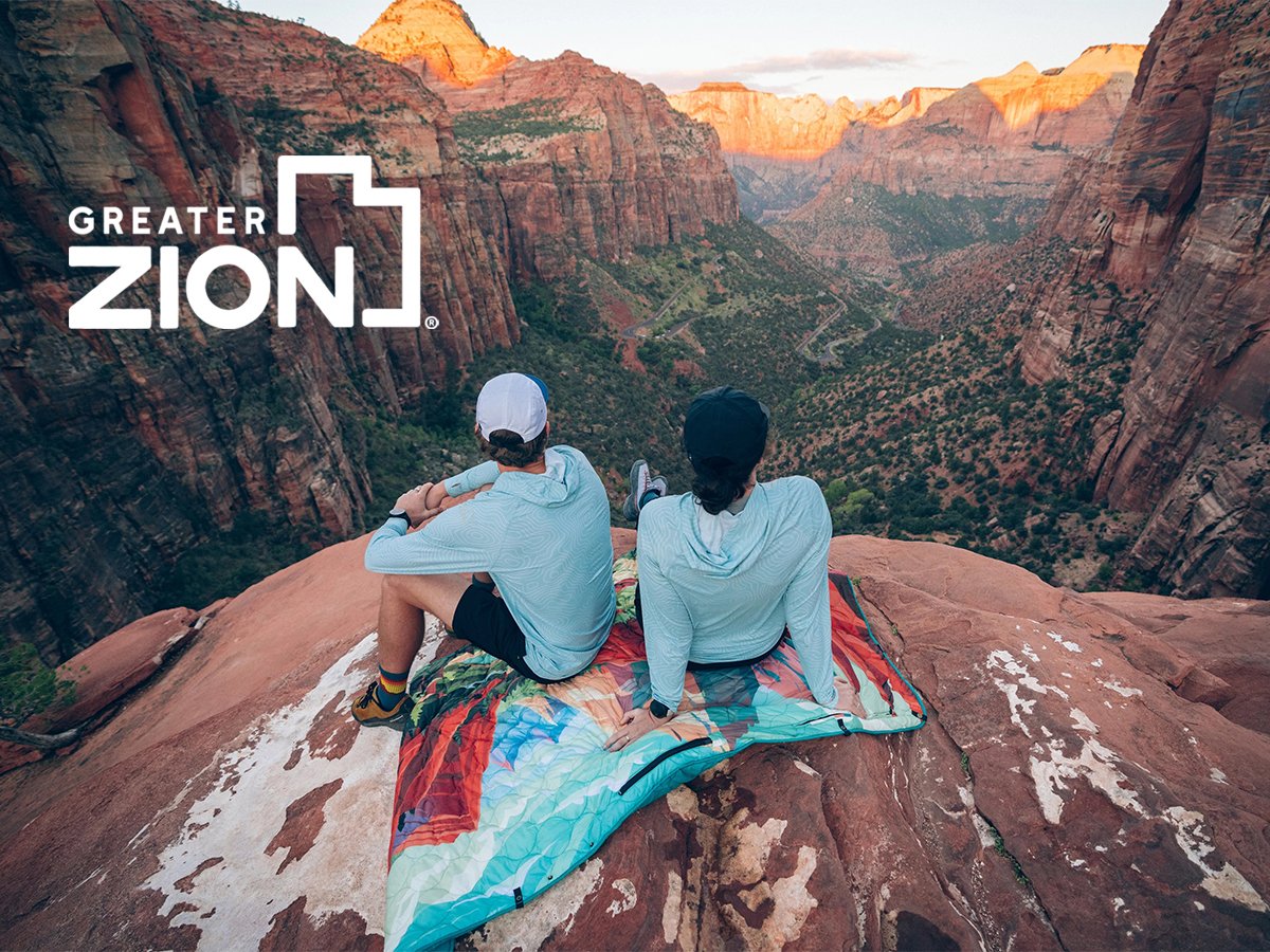 Two people sitting on a colorful blanket atop a red rock overlook, taking in a sweeping view of Zion Canyon at sunset with Greater Zion logo overlayed