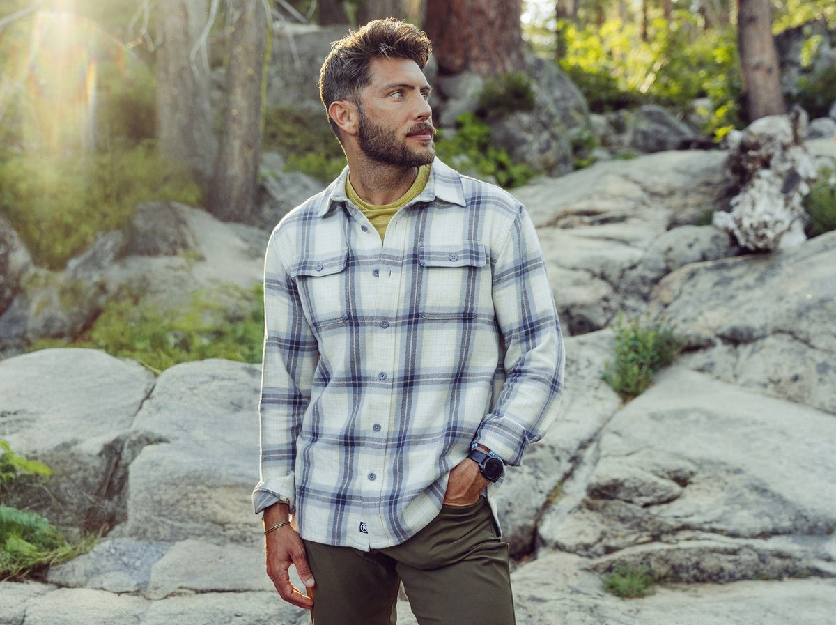 A person stands outdoors wearing a light-colored plaid button-up shirt over a yellow crew-neck top, with one hand in the pocket of olive green pants and a wristwatch visible.