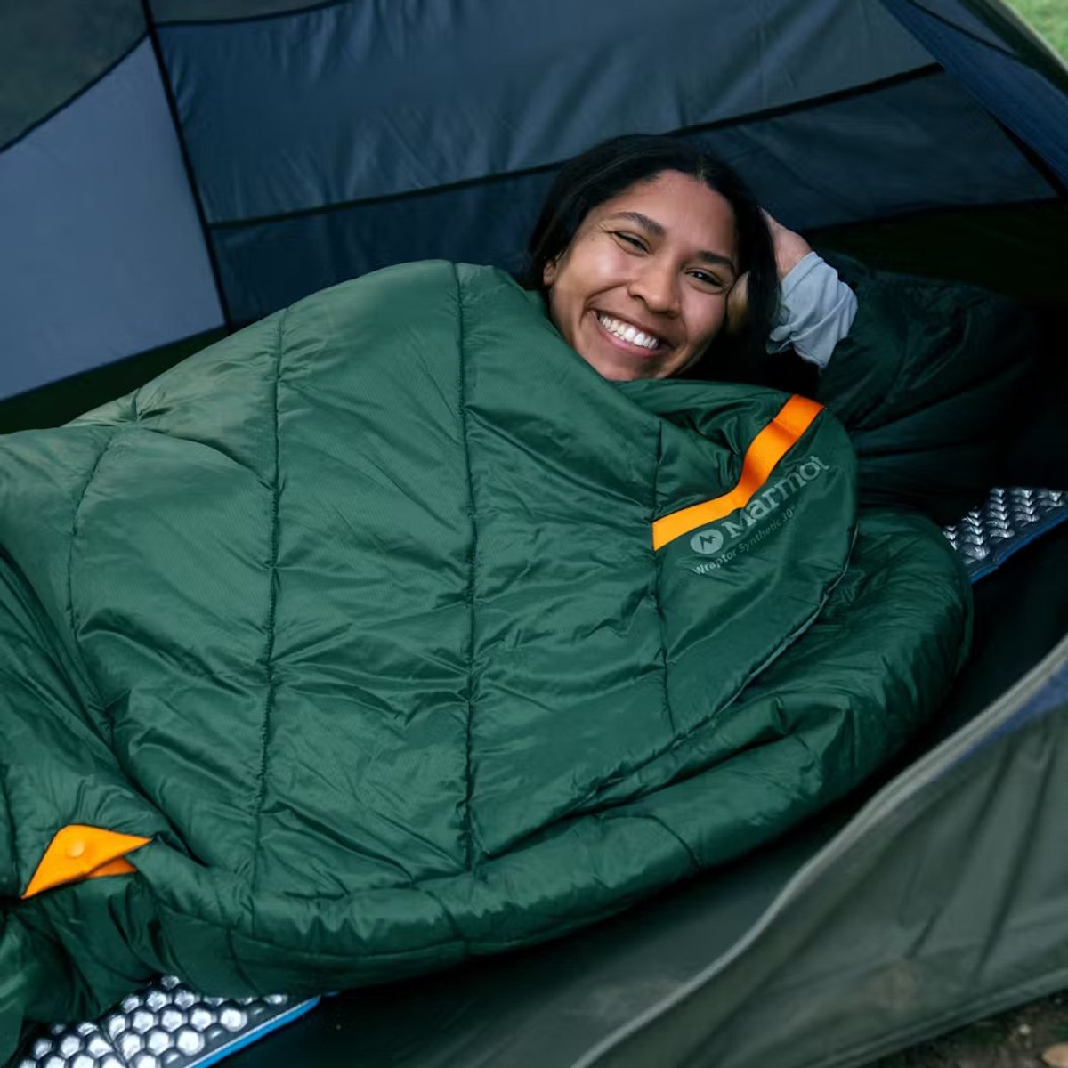 A woman happily lays in a tent, wrapped inside of the Marmot Wraptor sleeping bag.