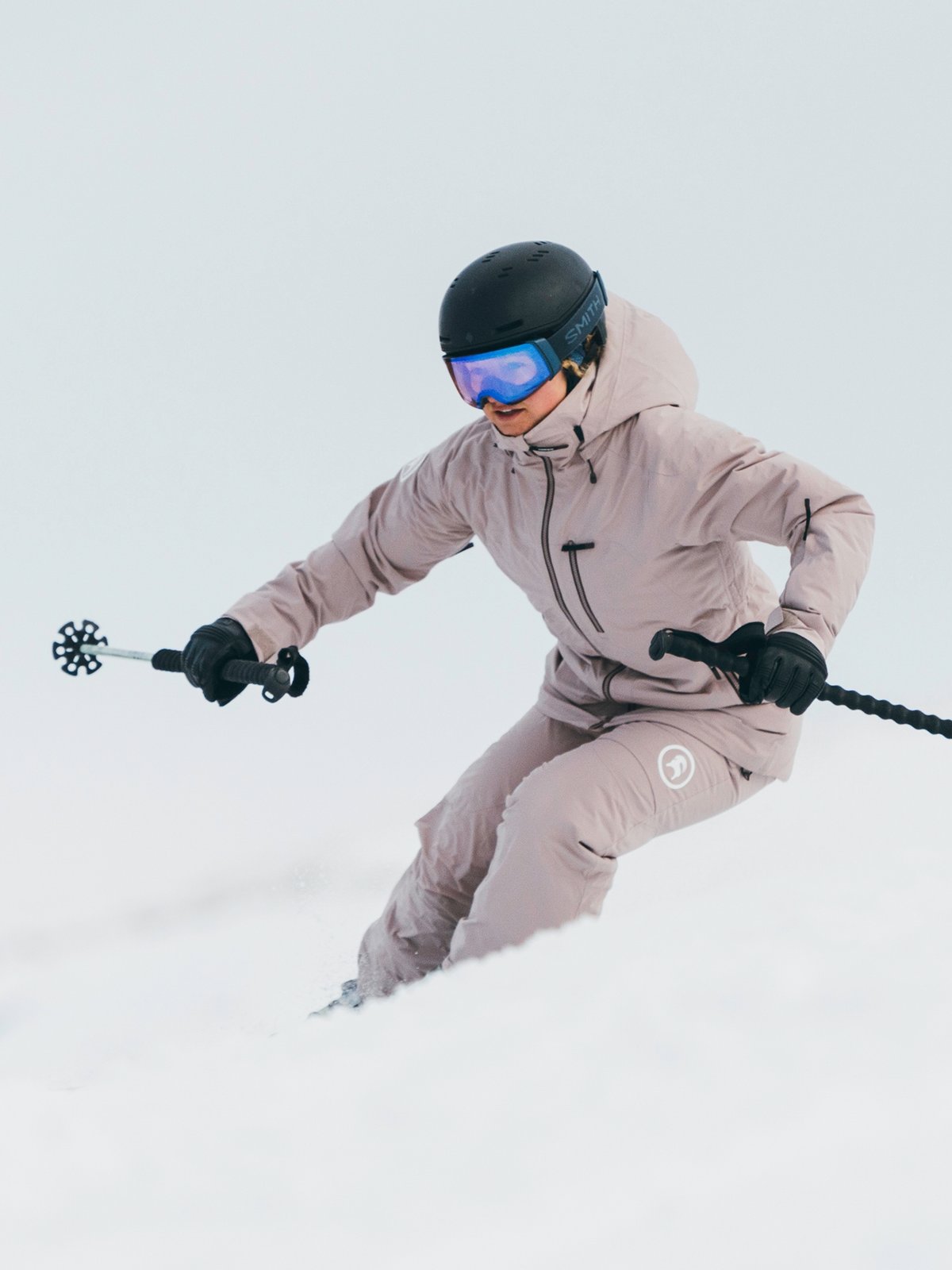 A skier dressed in a light mauve jacket and pants, black helmet, and blue goggles carves through fresh snow on a wintry slope while holding ski poles.