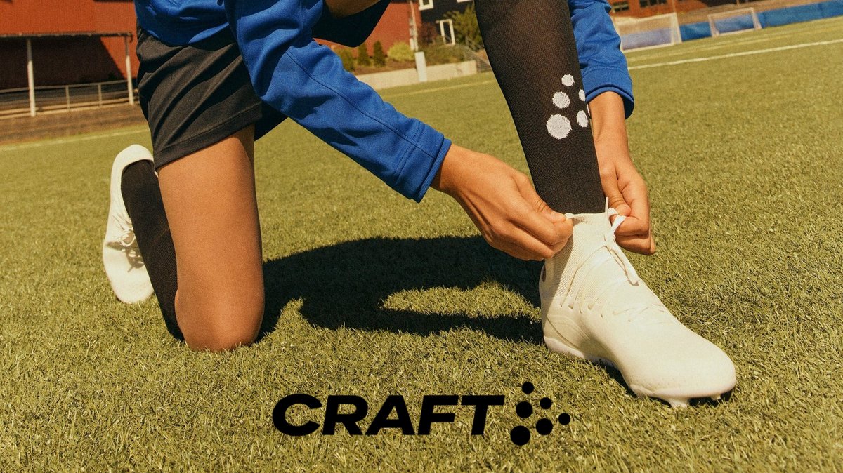 A person in blue athletic wear and white Craft socks adjusts their white sports shoe on a sunny outdoor field, showcasing performance footwear and athletic apparel.