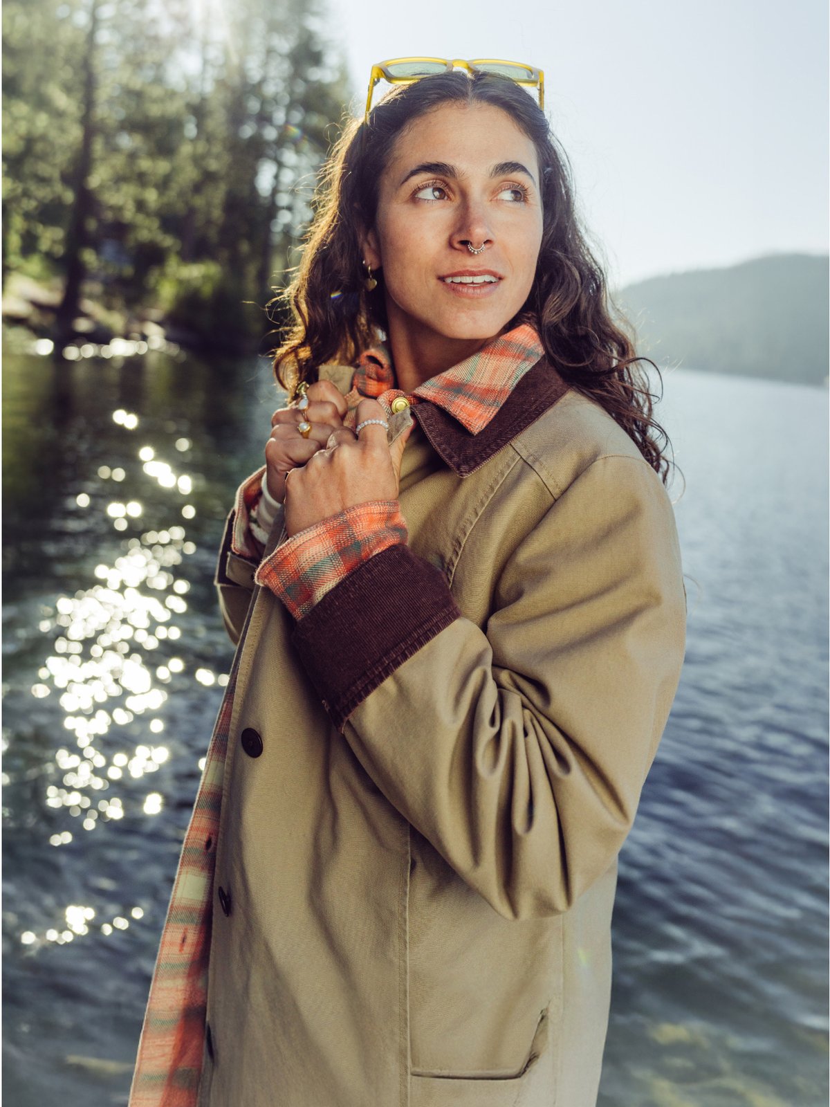 A woman with long curly hair stands by a sunlit lake wearing yellow sunglasses on her head, a tan coat with dark brown cuffs and collar, and an orange plaid shirt underneath.
